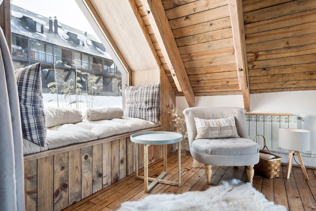 Une pièce chaleureuse avec un plafond en bois, un banc rembourré agrémenté de coussins, une petite table ronde, un fauteuil de couleur claire et un tapis blanc et moelleux sur le plancher en bois.