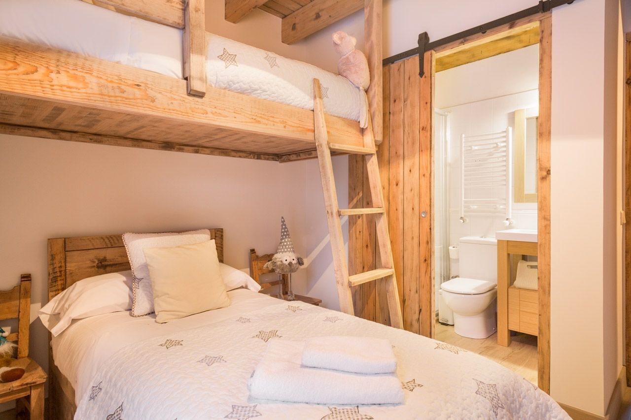 Een gezellige slaapkamer met een stapelbed, een houten ladder, een nachtkastje met een lamp, en een badkamer zichtbaar door een open deur.