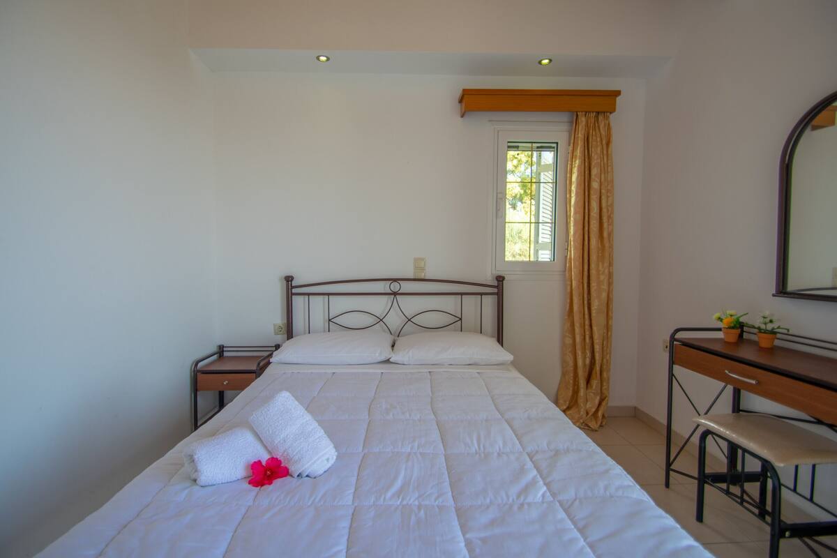A neatly made bed with a white comforter and a single red flower, a small wooden desk with a chair, a mirror, and a window with curtains.