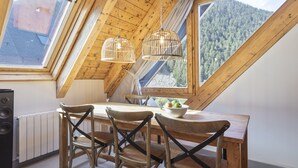 A dining area with a wooden table, chairs, and a bowl of fruit. There are two hanging light fixtures and a window with a view of a forested mountain. - Luderna - Val de Ruda C23 Barbacans (Baqueira)