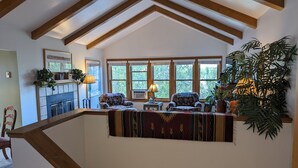 A cozy living room with a fireplace, two patterned armchairs, a coffee table, and large windows with a view of trees. - Durango Treehouse | Mountain Views | Hot Tub (Durango)