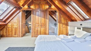Una habitación con techos altos, vigas de madera y tragaluces, una cama con sábanas blancas impecables, y un baño a la vista con lavabo y espejo.