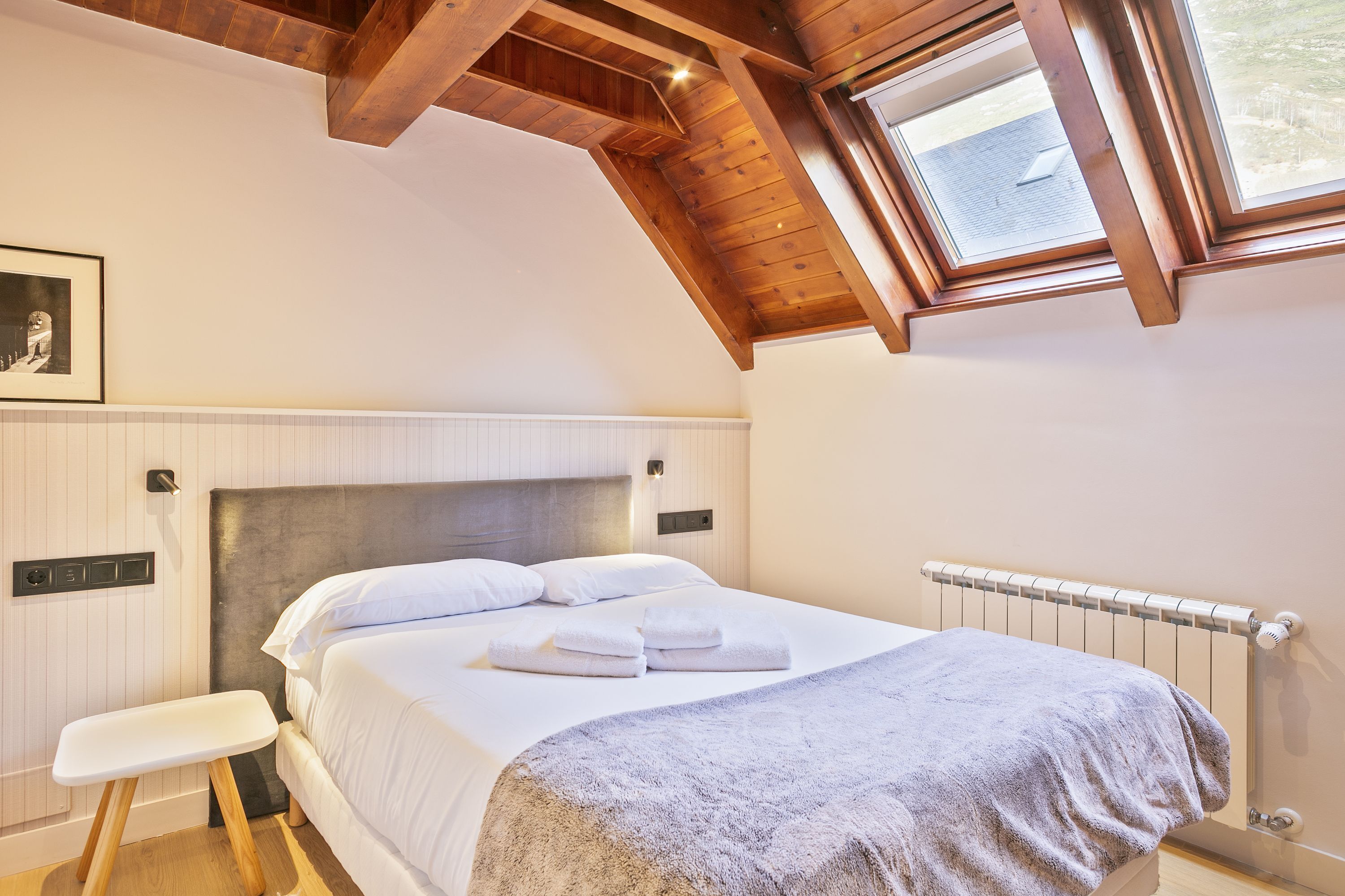 Un dormitorio con techo inclinado, una cama con ropa de cama blanca, una mesita de noche, un radiador y una ventana con vistas al exterior.