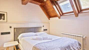 Un dormitorio con techo inclinado, una cama con ropa de cama blanca, una mesita de noche, un radiador y una ventana con vistas al exterior.