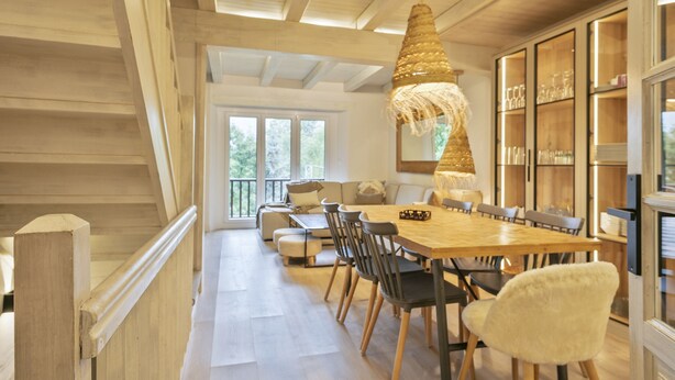 Un vaste espace dédié à la salle à manger, avec une table et des chaises en bois, un luminaire suspendu et une vue sur l’extérieur grâce à de grandes fenêtres.