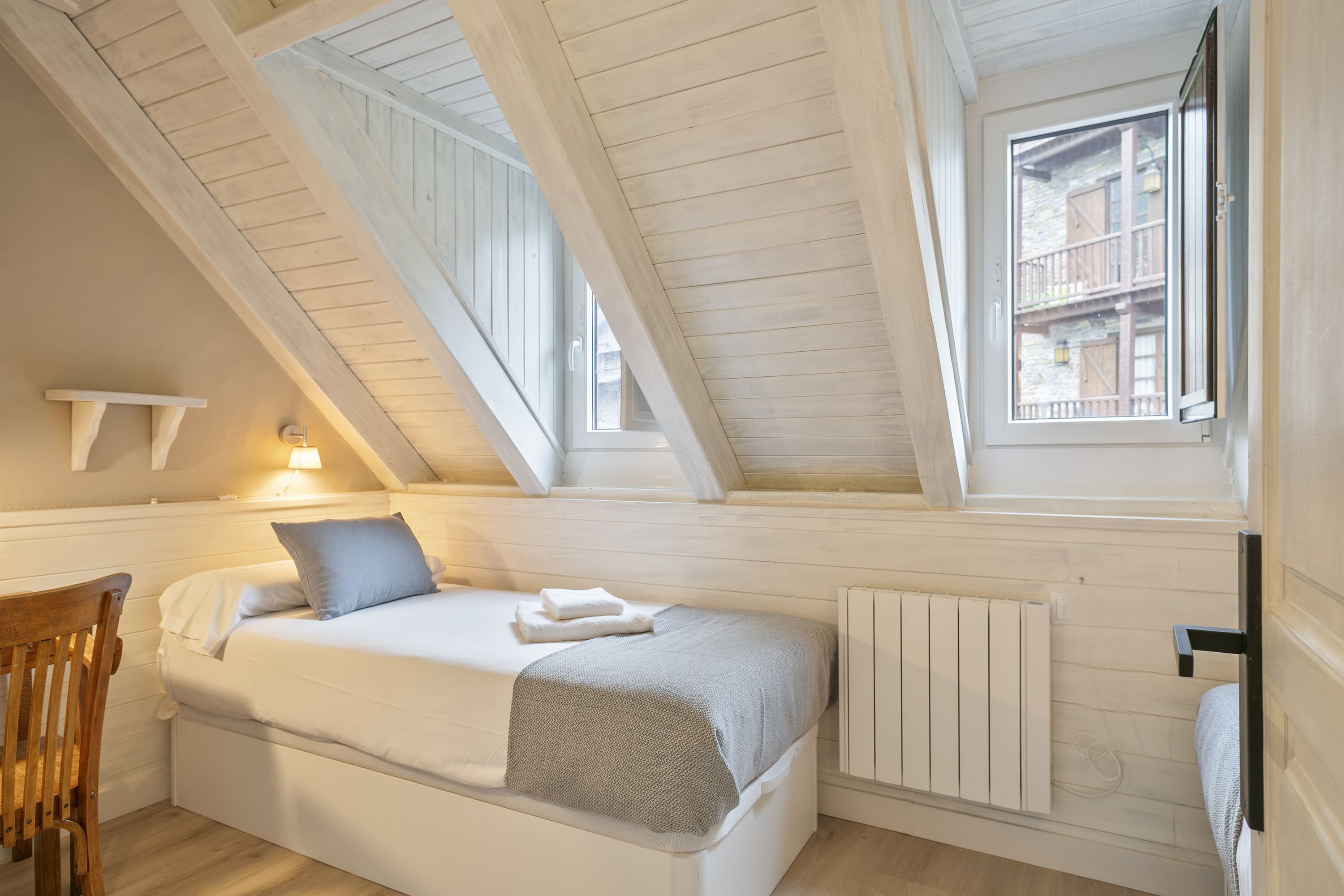 A bedroom with a sloped ceiling, a bed with a white headboard, a wooden chair, and a window with a view of buildings.