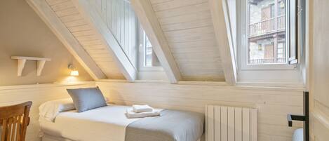 A bedroom with a sloped ceiling, a bed with a white headboard, a wooden chair, and a window with a view of buildings.