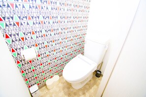 A bathroom with a white toilet, a toilet brush, and a toilet paper holder. The walls are tiled with a geometric pattern. - Grande Shagi GS505 (Osaka)