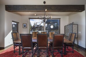 A dining area with a wooden table and six chairs, a chandelier, and a view of a house through large windows. - Newly built Luxury Home, 5-Star Finishes, SHUTTLE to slopes, backs to Breckenridge Nordic Center! (Breckenridge)