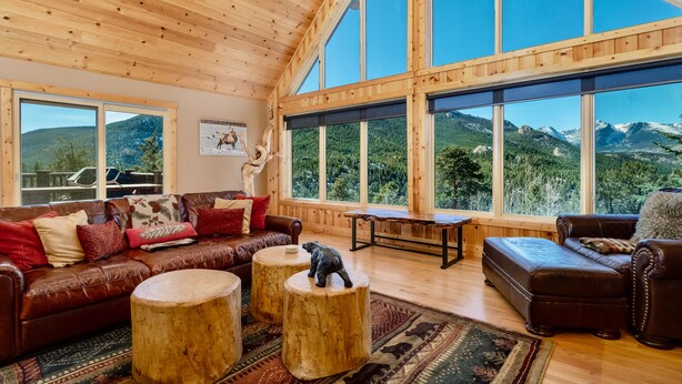 Ein großzügiges Wohnzimmer mit einer hohen Holzbalkendecke, einem braunen Ledersofa, runden Holzschemeln und Bergblick durch große Fenster.