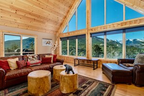 A spacious living room with a large wooden ceiling, a brown leather sofa, round wooden stools, and a view of mountains through large windows. - Awestruck Luxury Vacation Home at Windcliff (Estes Park)