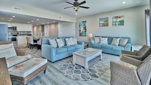 A spacious living room with a blue sofa set, a coffee table, and wicker chairs. There is a dining area with a table and chairs, and a kitchen in the background. - PierWatch I 101 | Oceanfront Six Bedroom Condo (North Myrtle Beach)