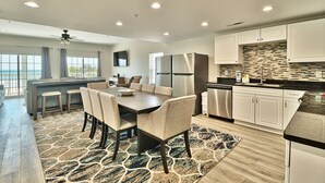 A modern kitchen with white cabinets, a stainless steel refrigerator, and a dining area with a large table and chairs. - PierWatch I 101 | Oceanfront Six Bedroom Condo (North Myrtle Beach)