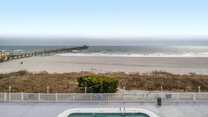 A beach view with a wooden pier extending into the ocean, a white fence, and a small bush in the foreground. - PierWatch I 103 | Oceanfront Six Bedroom Condo (North Myrtle Beach)