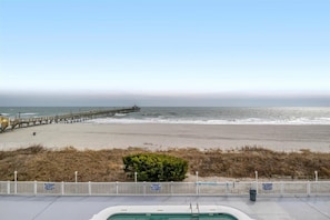 Blick auf einen Strand mit einer Holzstege, der sich ins Meer erstreckt, einem weißen Zaun und einem kleinen Busch im Vordergrund.