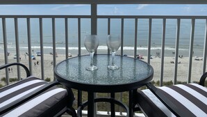 A balcony with a glass table and two wine glasses, overlooking a beach with people and umbrellas. - Ocean Front Resort in North Myrtle Beach,  Sleeps 6, Directly on the Beach! (North Myrtle Beach)