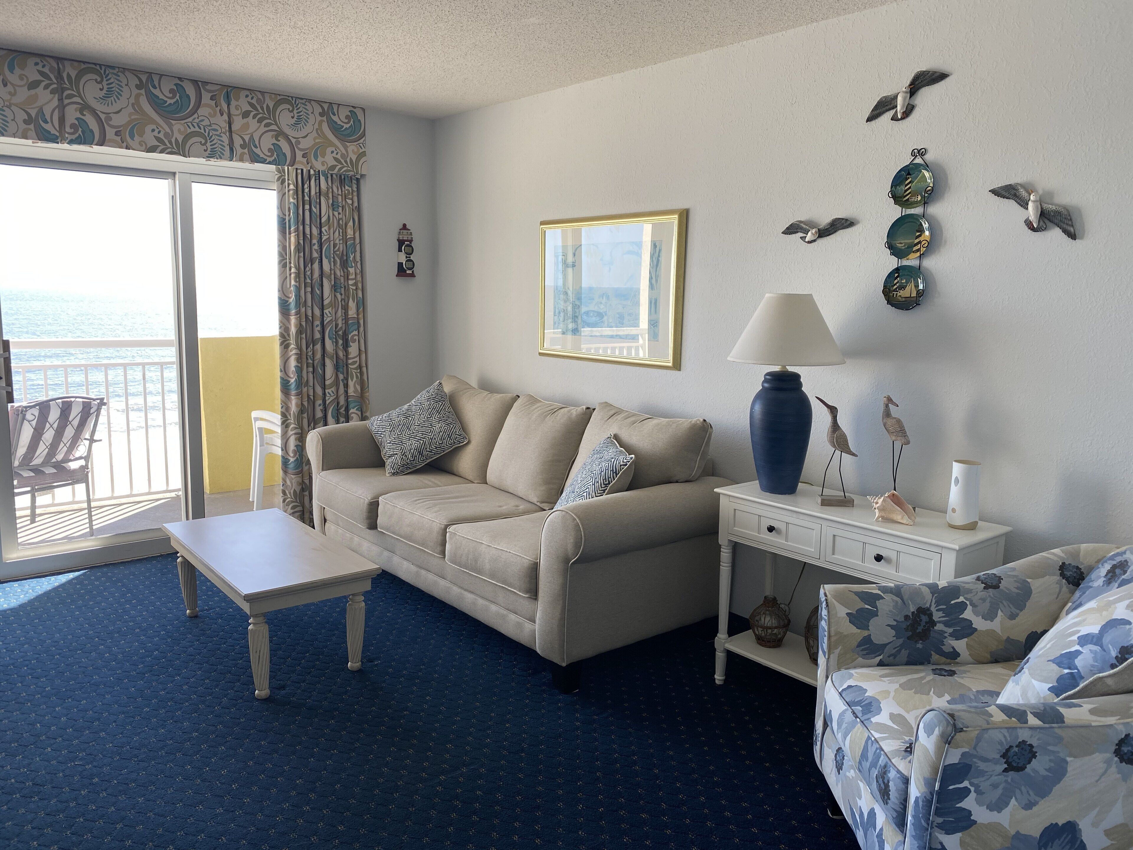 A living room with a sofa, a coffee table, and a side table with a lamp. There is a window with curtains, a framed picture on the wall, and decorative wall art.