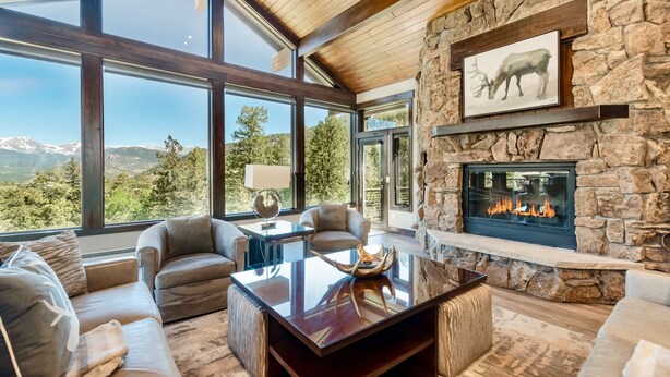 A spacious living room with a stone fireplace, large windows offering mountain views, and comfortable seating. - Hyler Mountain Lodge Vacation Home at Windcliff (Estes Park)