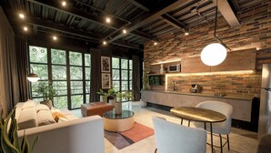 A modern living room with a sofa, coffee table, and chairs, a kitchen with a brick wall, and large windows with natural light. - Modern 1BR Loft in the middle of Provenza (Medellín)