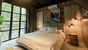 A modern bedroom with a large bed, a view of trees through a window, and a brick wall. - Modern 1BR Loft in the middle of Provenza (Medellín)