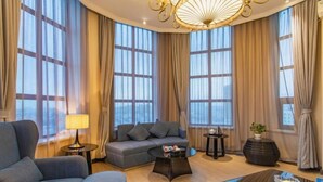 Una sala de estar moderna con sofá, sillón y una mesa de centro, ventanas amplias con cortinas y una araña.