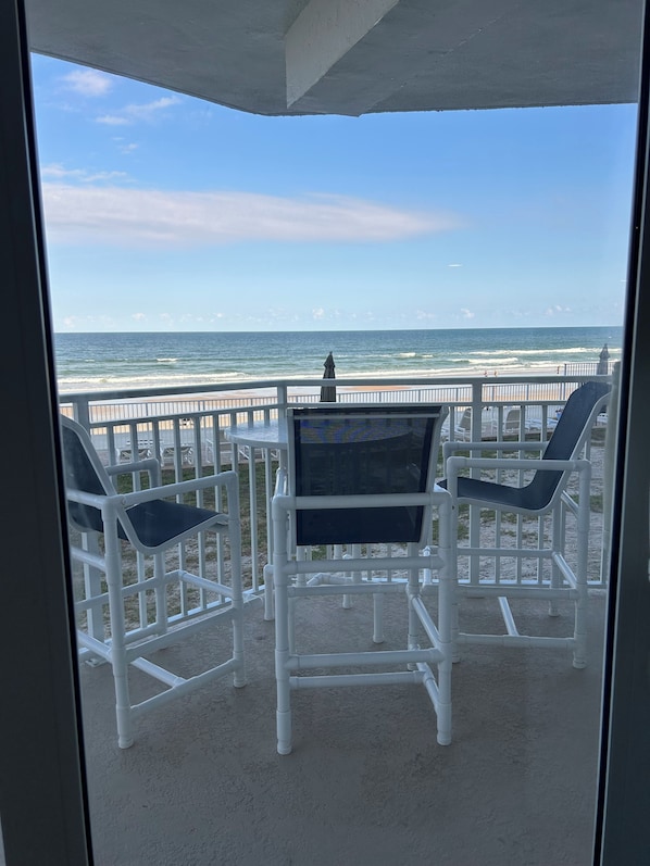 A balcony with white chairs and a table, overlooking a beach with a railing and ocean view. - OCEANFRONT 3BD-3B Condo - ground floor. (Daytona Beach)