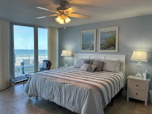 A bedroom with a bed, two bedside lamps, a nightstand, a chair, and a balcony with a view of the ocean.