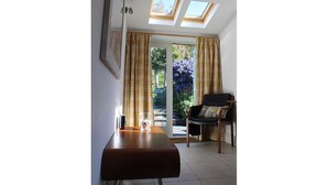 A room with a wooden table, a chair, and a glass of water. There are two skylights above a sliding glass door leading to an outdoor area with plants.