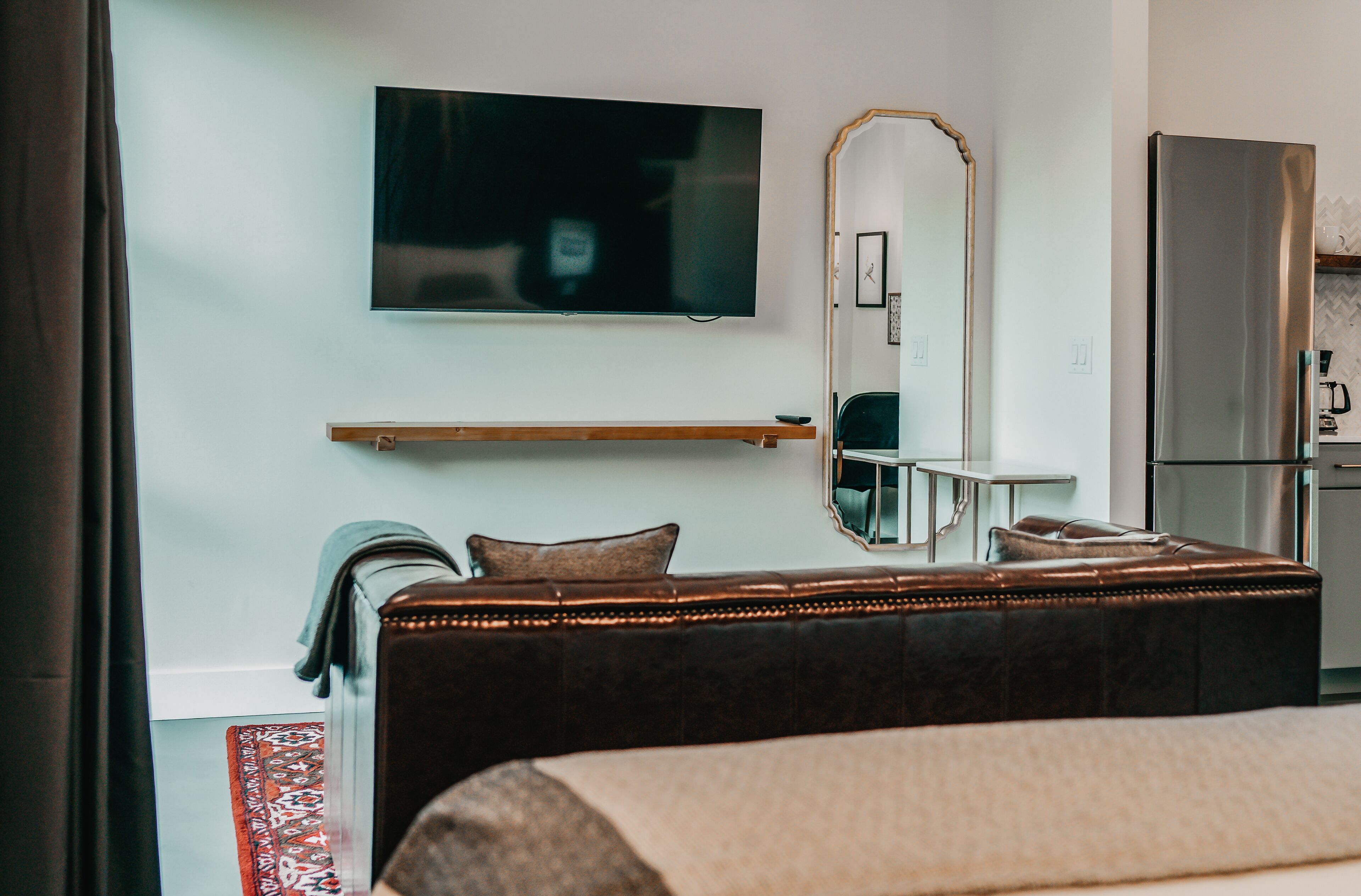 A hotel room with a bed, a television mounted on the wall, a mirror, a small table, and a refrigerator.