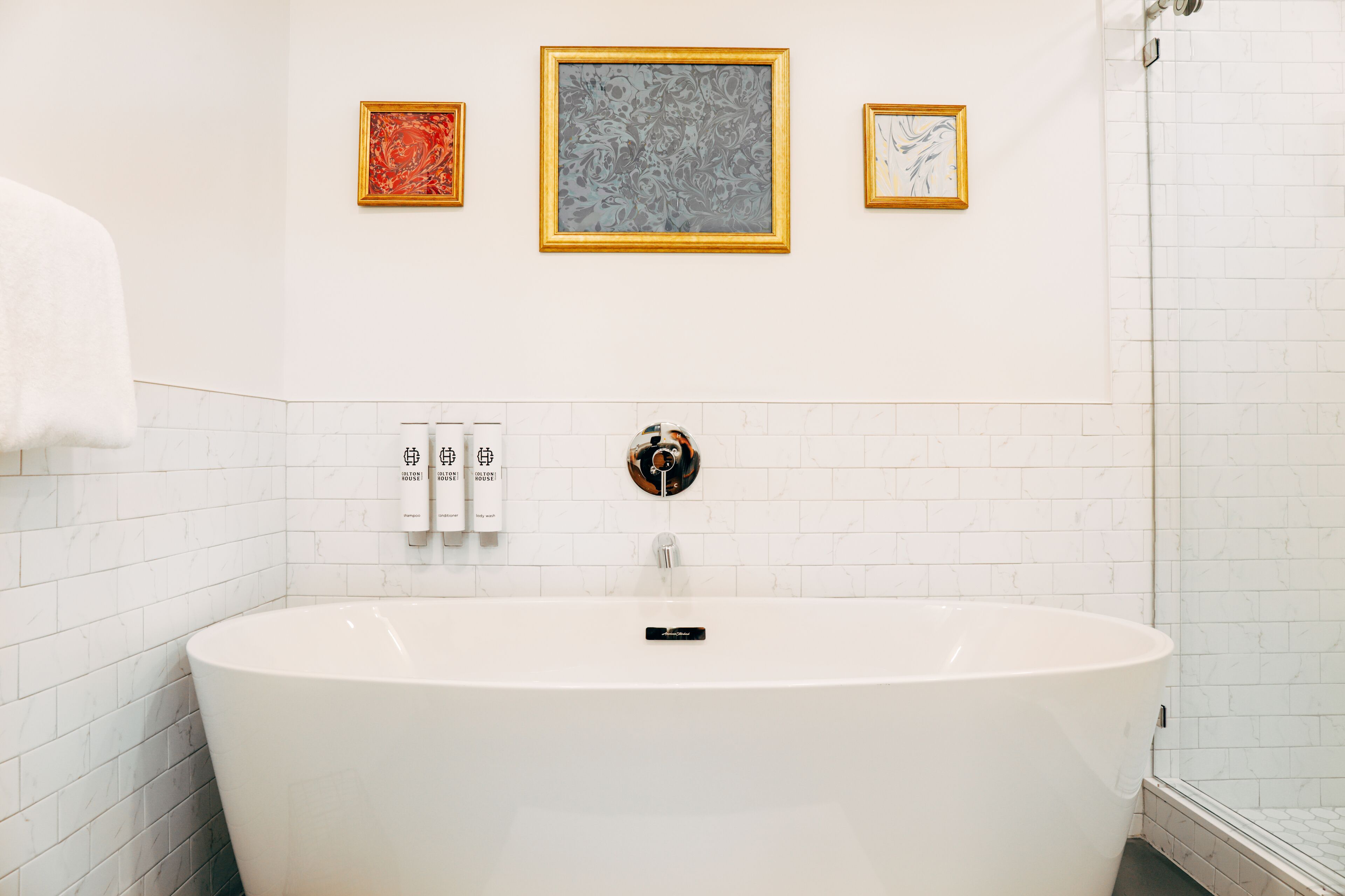 A modern bathroom with a freestanding bathtub, white tiled walls, and three framed artworks above the tub.