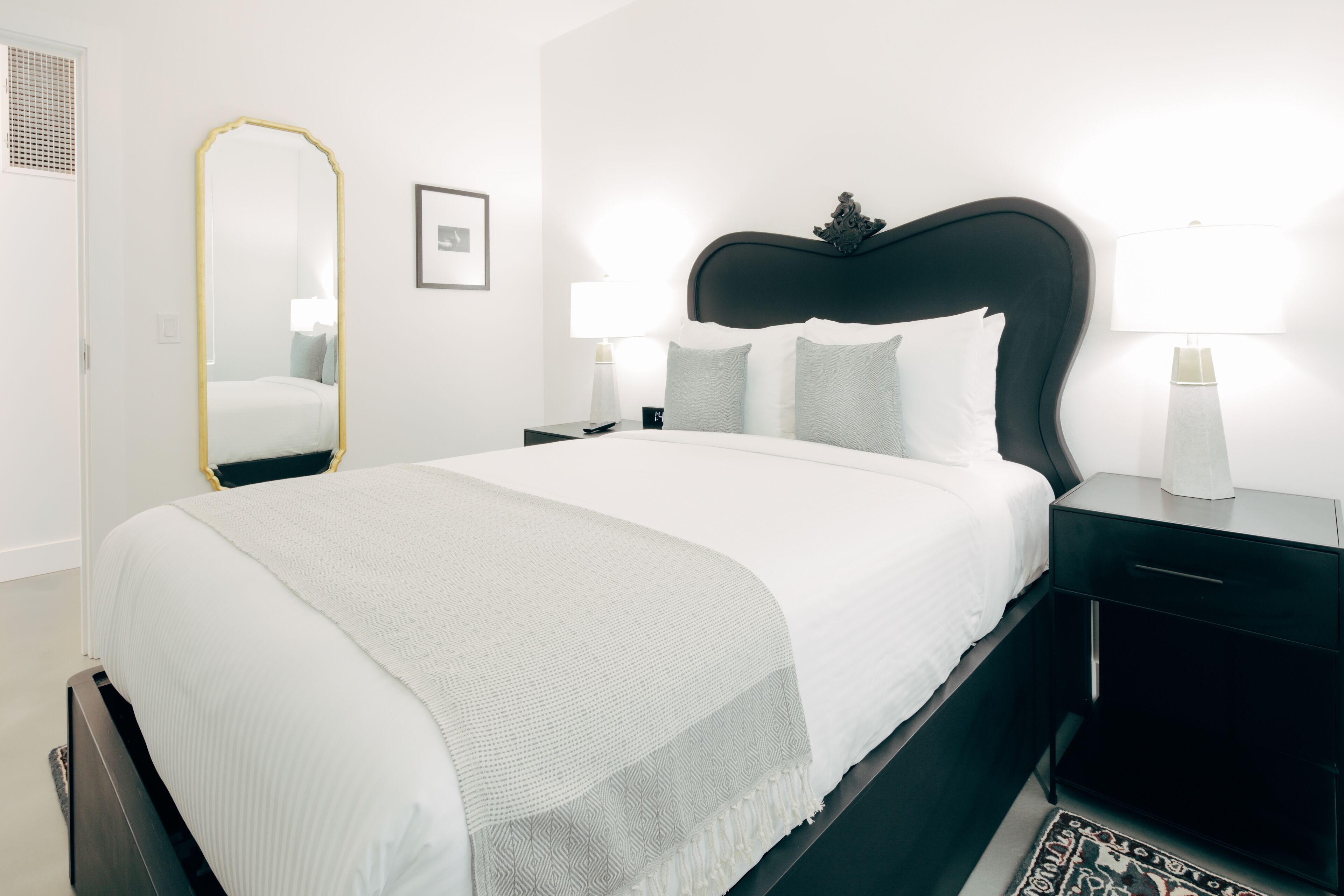 A neatly arranged bedroom with a large bed, a bedside table with a lamp, a mirror, and a framed picture on the wall.