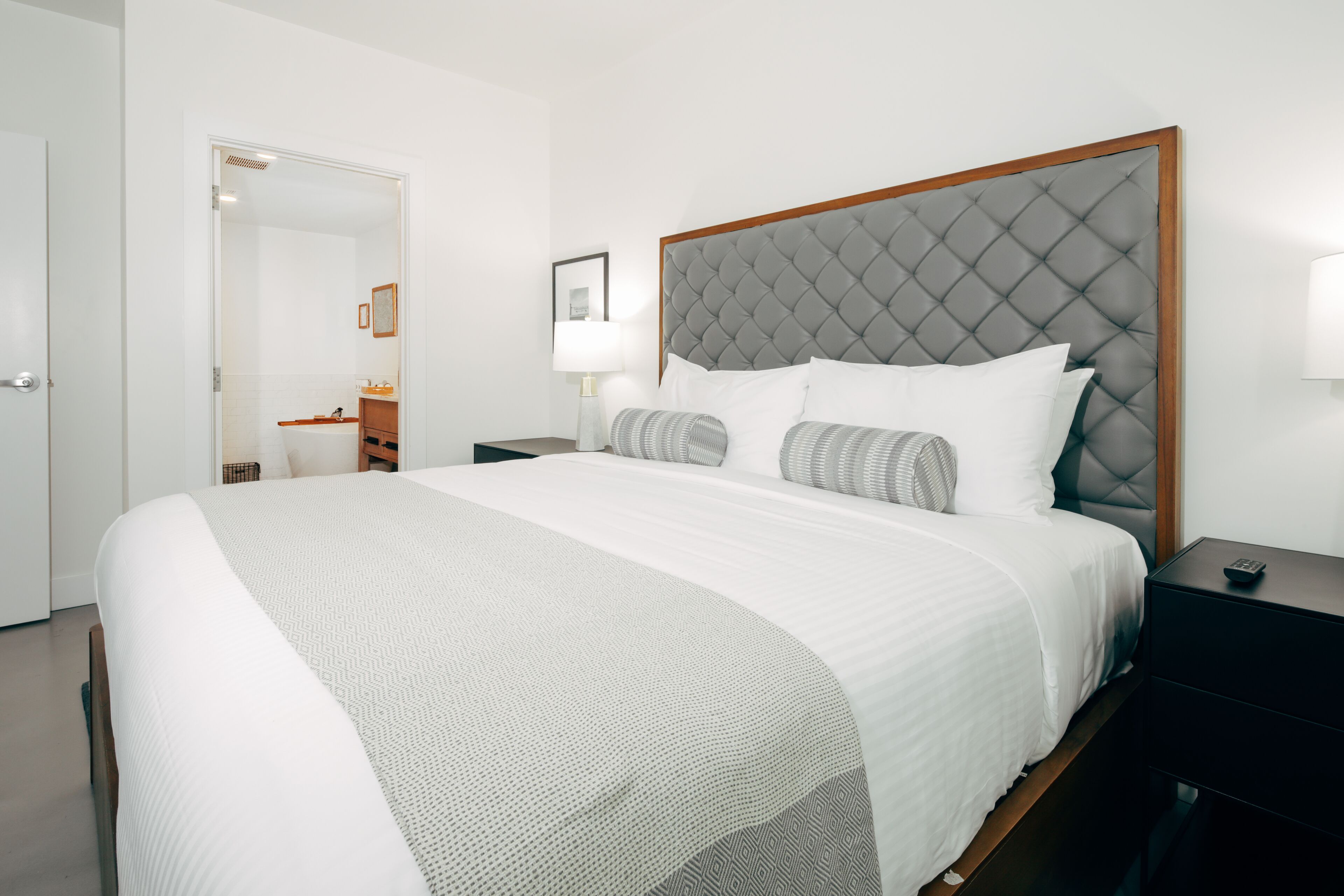 A neatly made bed with a grey tufted headboard, white bedding, and a bedside table with a lamp.