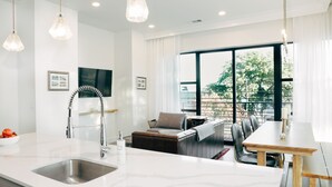 Een moderne keuken met een wit aanrecht, een roestvrijstalen spoelbak en hanglampen. Er is een eethoek met een houten tafel en stoelen, en een bank met uitzicht op de omgeving door grote ramen.