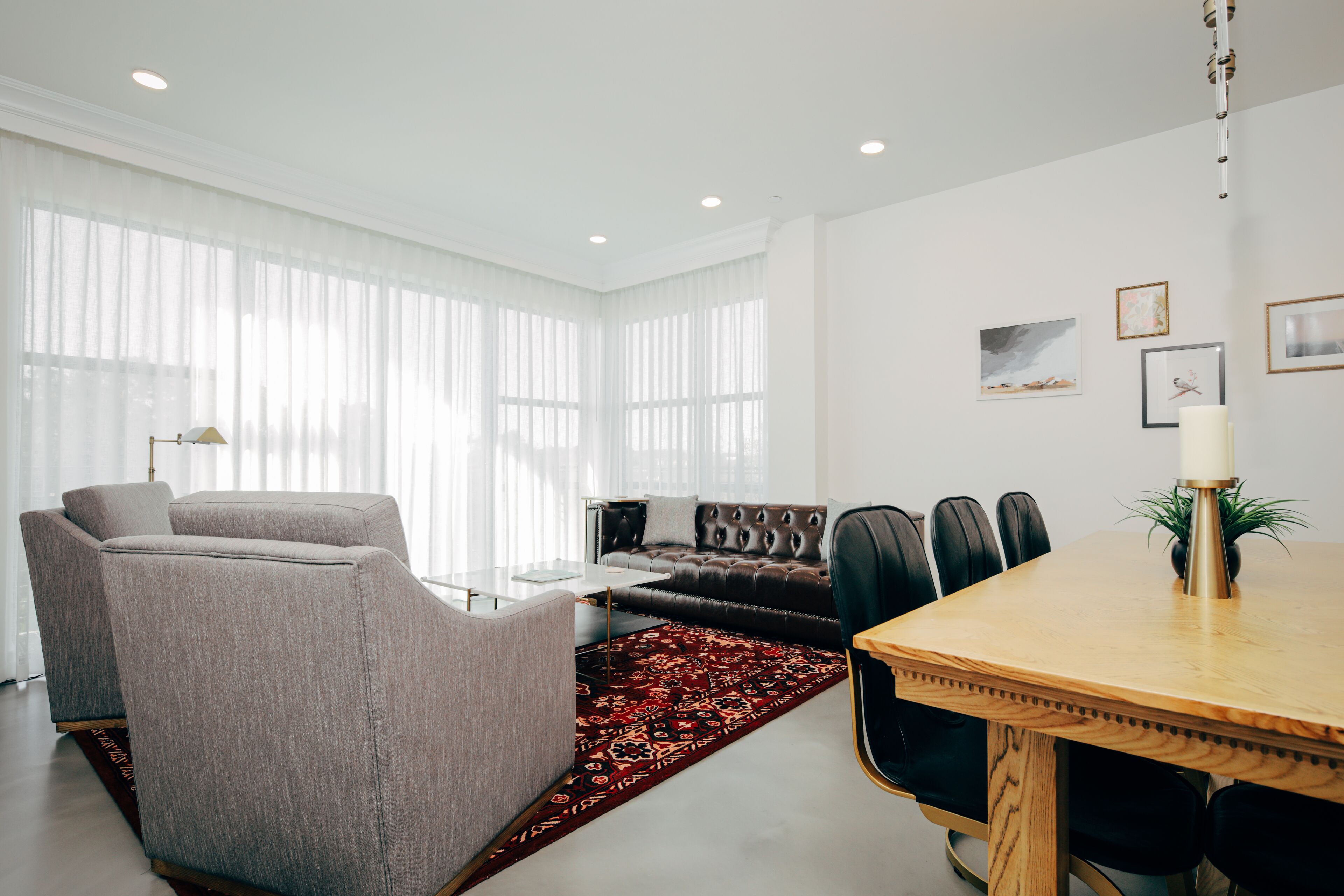 A modern living room with a sofa, chairs, a dining table, and a large window with sheer curtains.