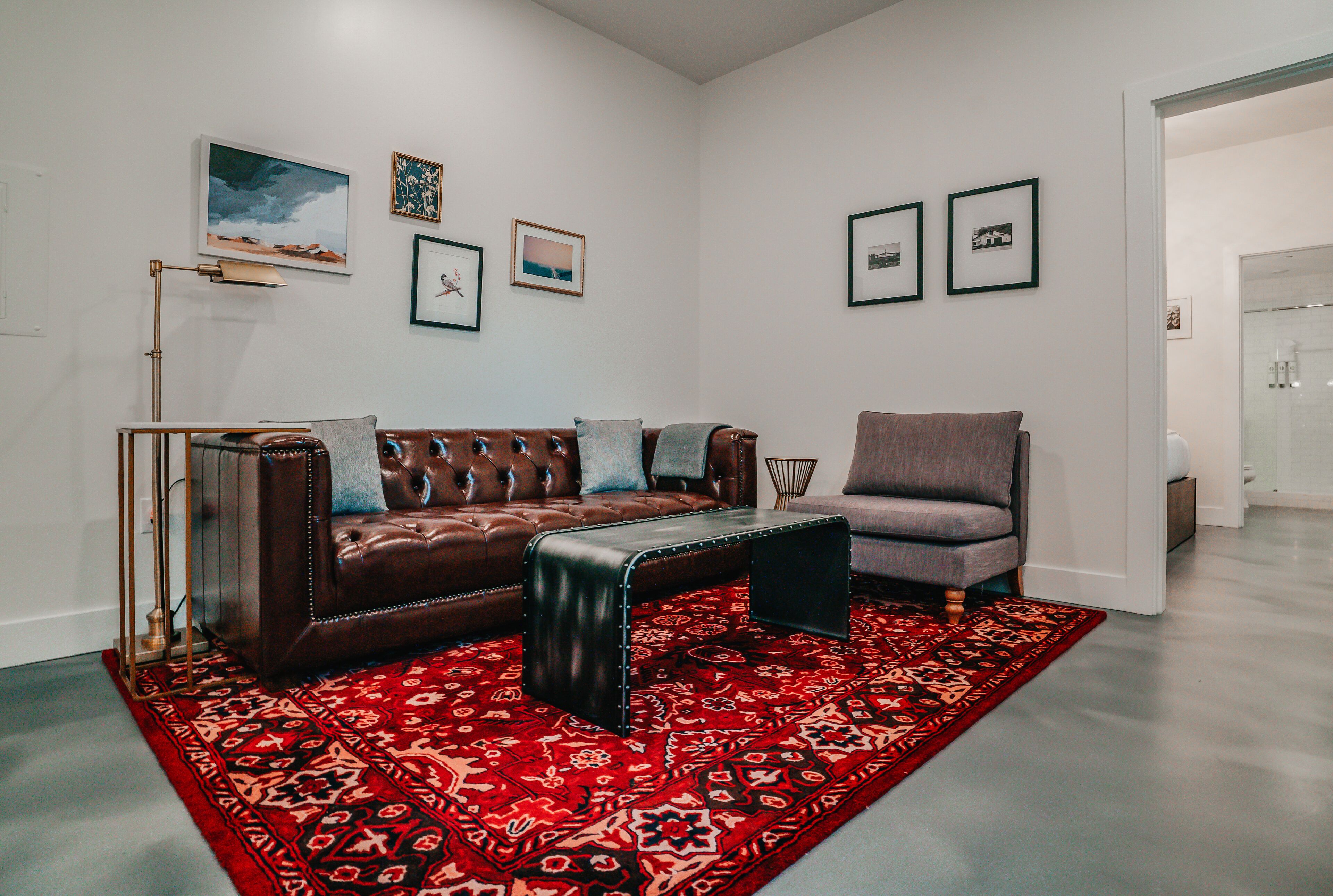A living room with a brown leather sofa, a grey armchair, and a red patterned rug. There are framed pictures on the walls.