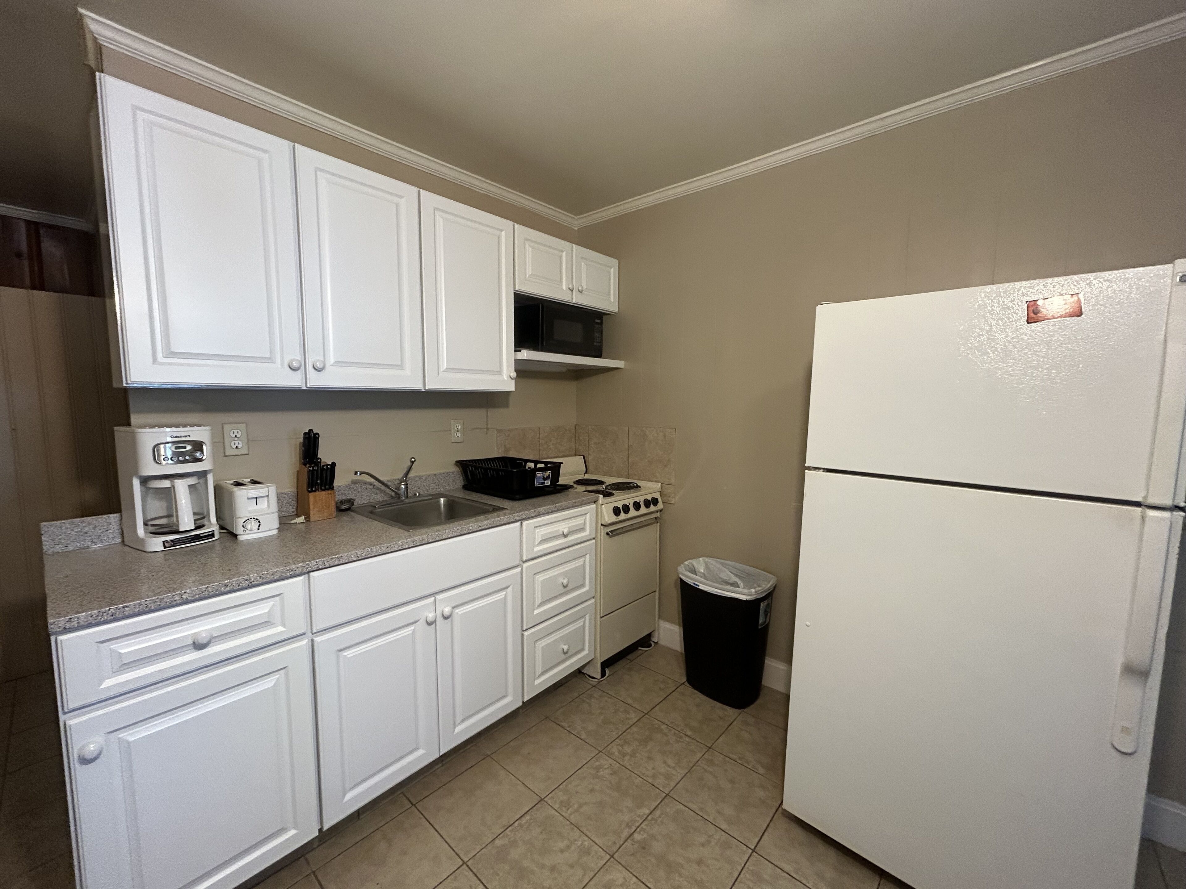 A kitchen with white cabinets, a stove, a sink, a coffee maker, and a trash can.