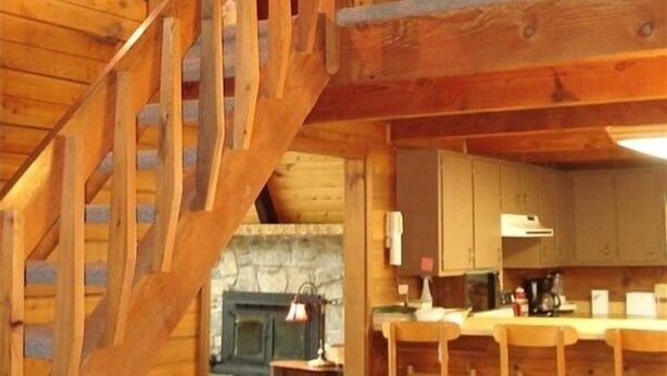 A wooden interior with a staircase leading to an upper level, a kitchen area with bar stools, and a stone fireplace. - Spacious Mountain Cabin in Gated Community (Triplett)