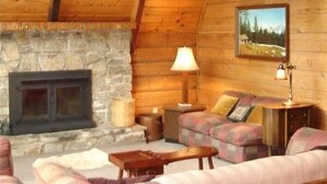 A cozy log cabin interior with a stone fireplace, plaid furniture, and a painting on the wall. - Spacious Mountain Cabin in Gated Community (Triplett)