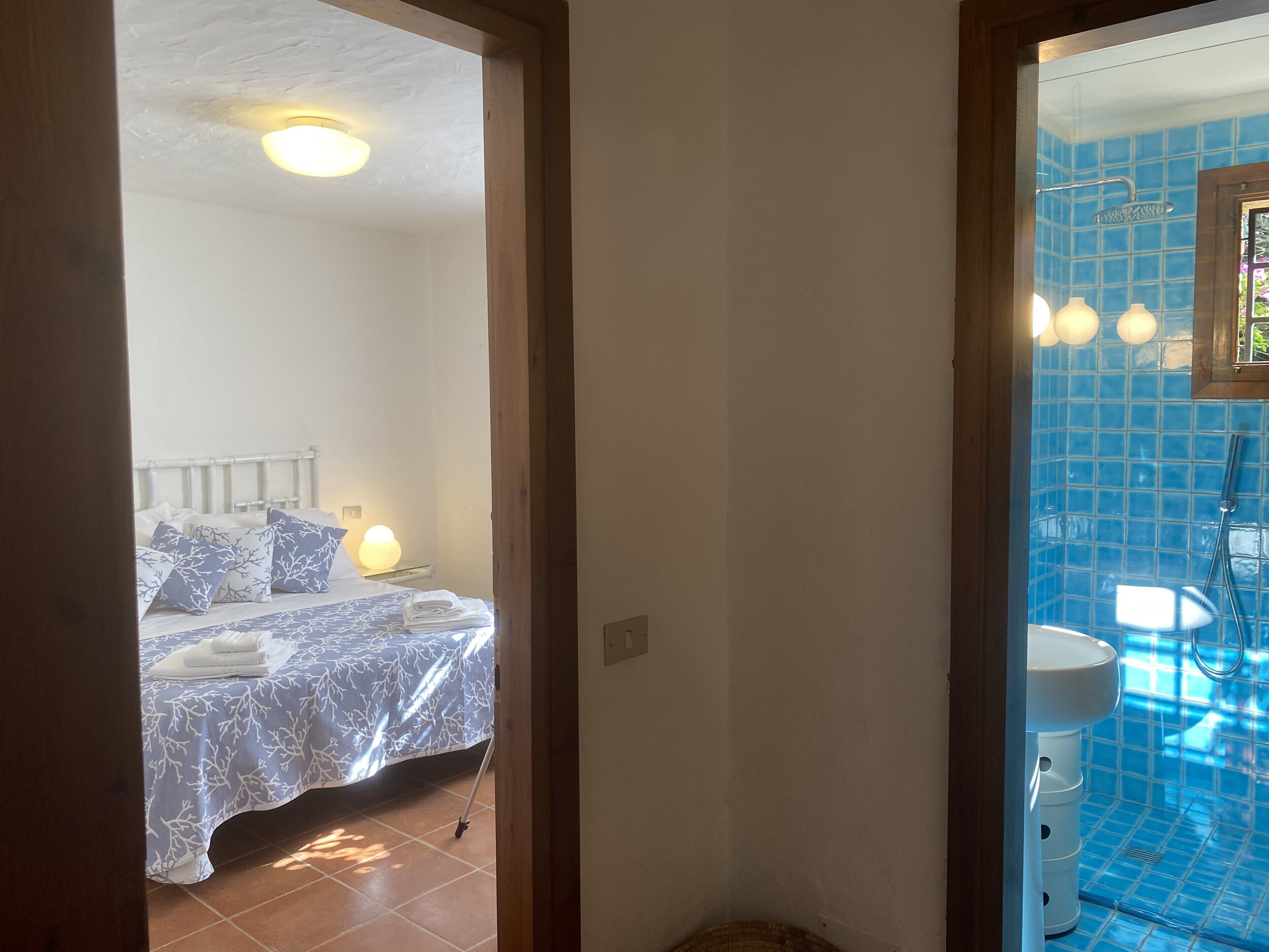 A bedroom with a bed, pillows, a bedside lamp, and a bathroom with a sink and blue-tiled walls.