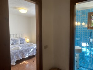 Ein Schlafzimmer mit Bett, Kissen, einer Nachttischlampe und einem Badezimmer mit Waschbecken und blauen Fliesen.