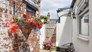 Eine Backsteinmauer mit einem hängenden Blumenkorb, eine weiße Mauer mit einem Fenster sowie eine Terrasse mit Tisch und Stühlen.