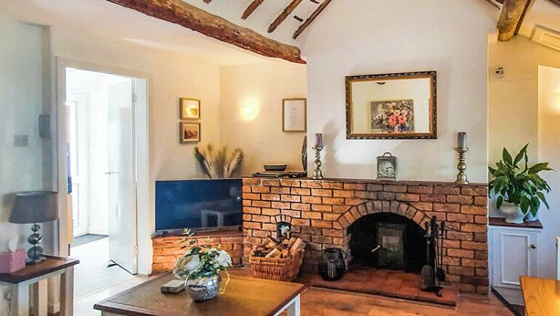 Ein gemütliches Wohnzimmer mit Kamin aus Ziegelsteinen, Holzbalken an der Decke, einem Fernseher und einer Topfpflanze.