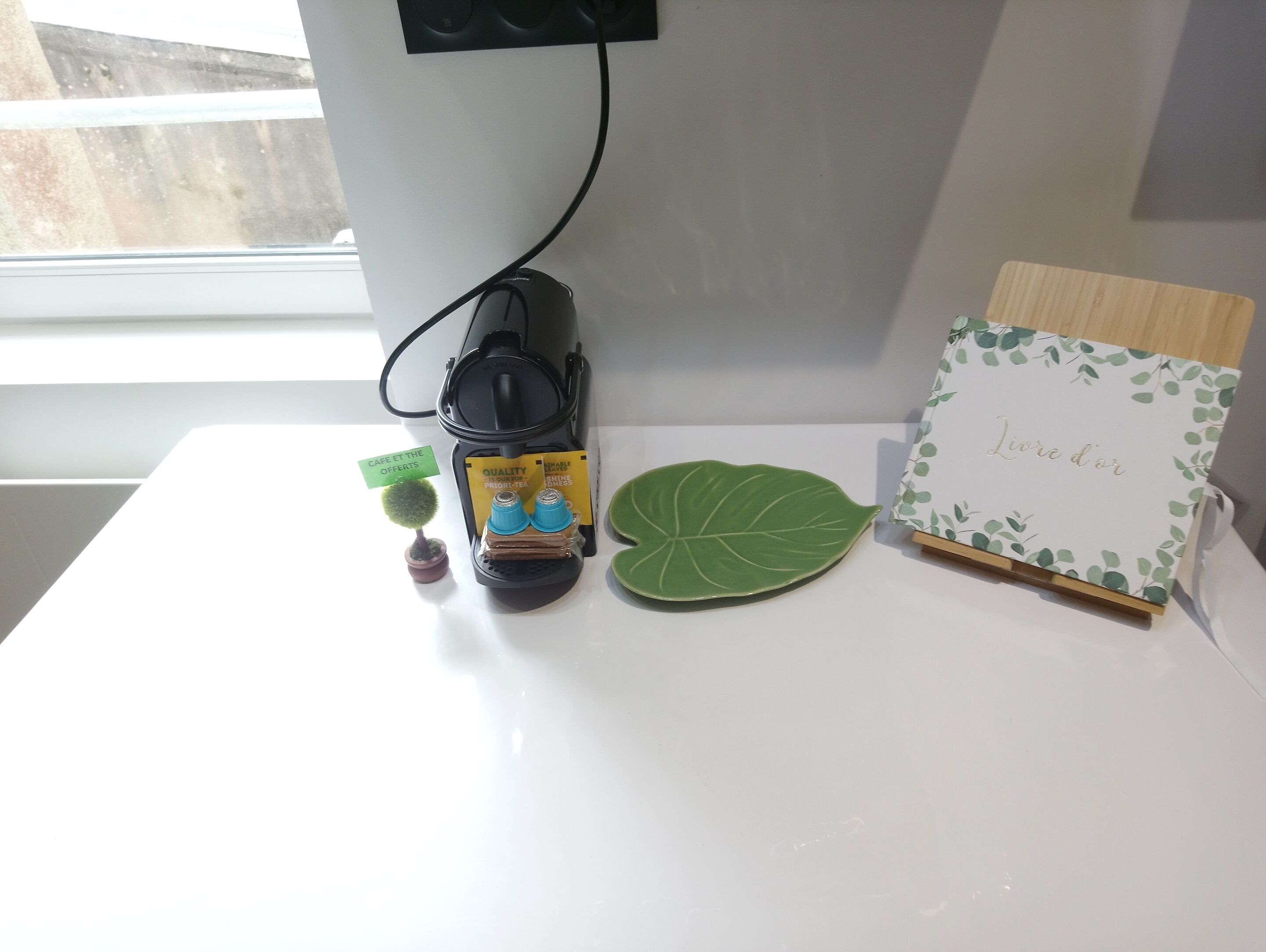 Une table avec une machine à café, une assiette en forme de feuille verte, une petite plante en pot et une carte décorative.