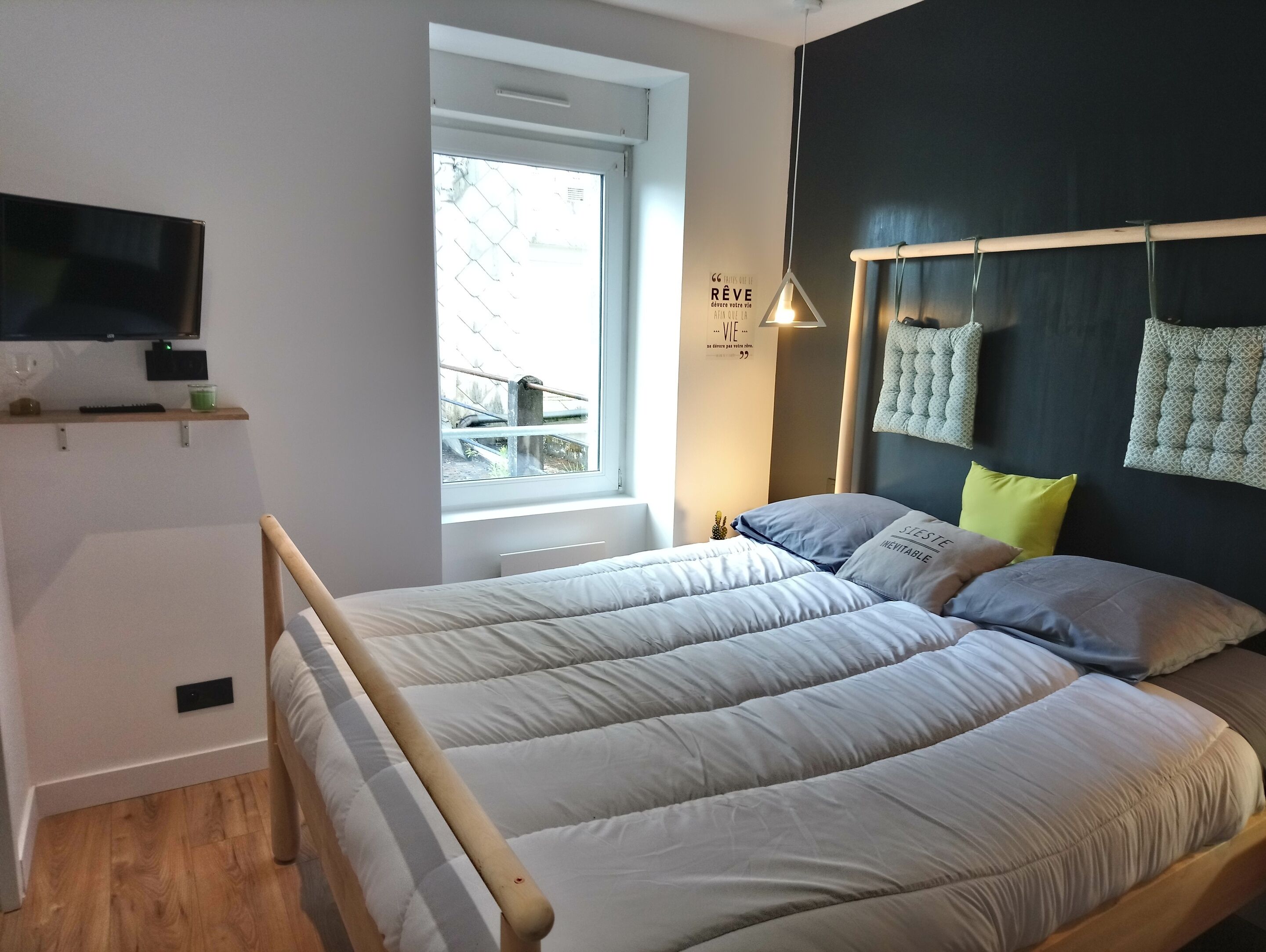 Un dormitorio con una cama, televisión montada en la pared, una ventana y una lámpara de noche.