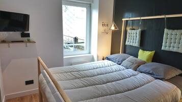 Un dormitorio con una cama, televisión montada en la pared, una ventana y una lámpara de noche.