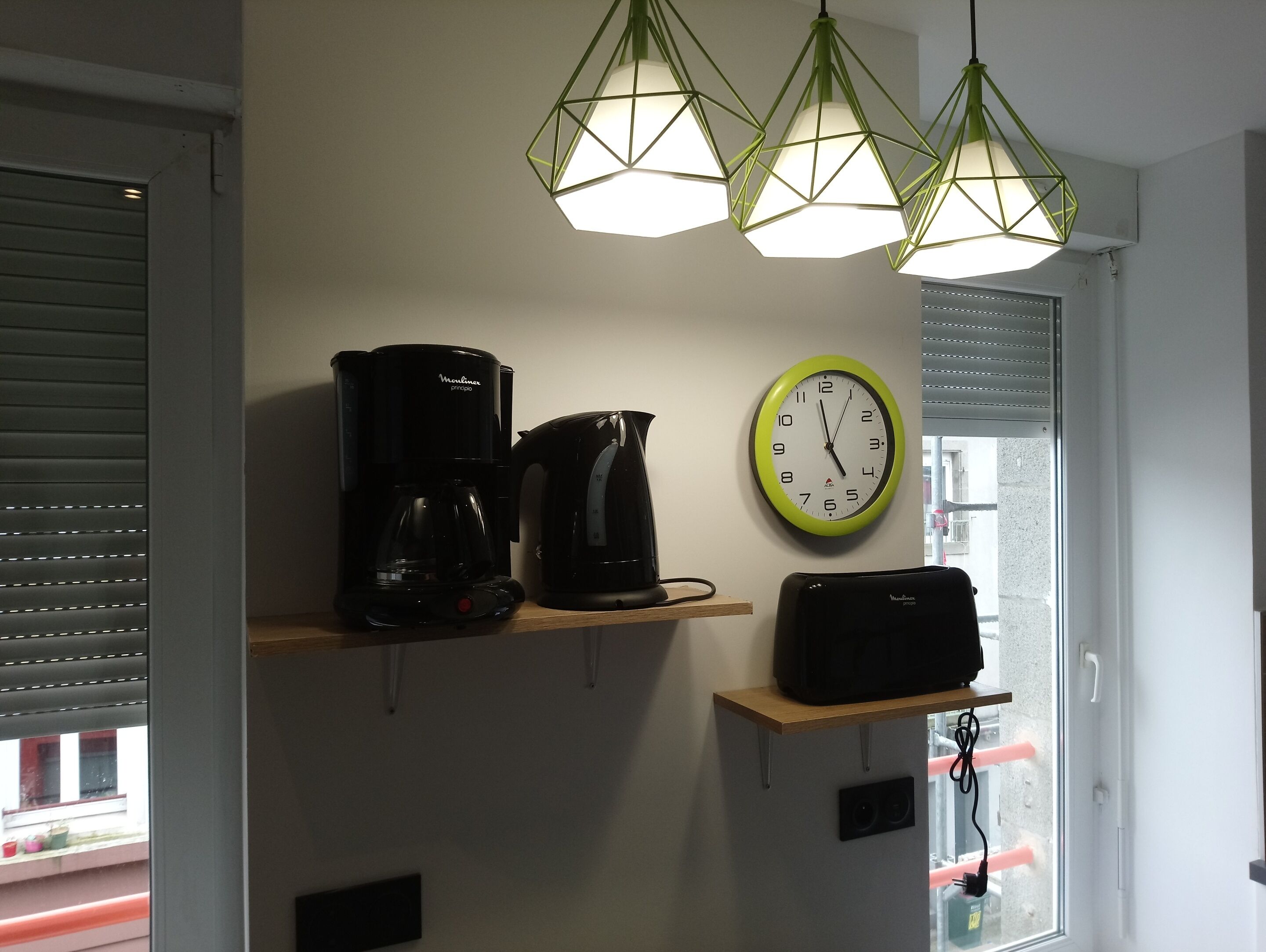 A kitchen with a coffee maker, kettle, and microwave on shelves, a clock, and a window with blinds.