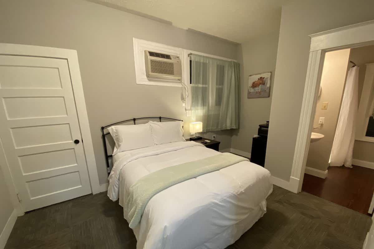 A bedroom with a bed, a nightstand, a lamp, a window with curtains, and a door.