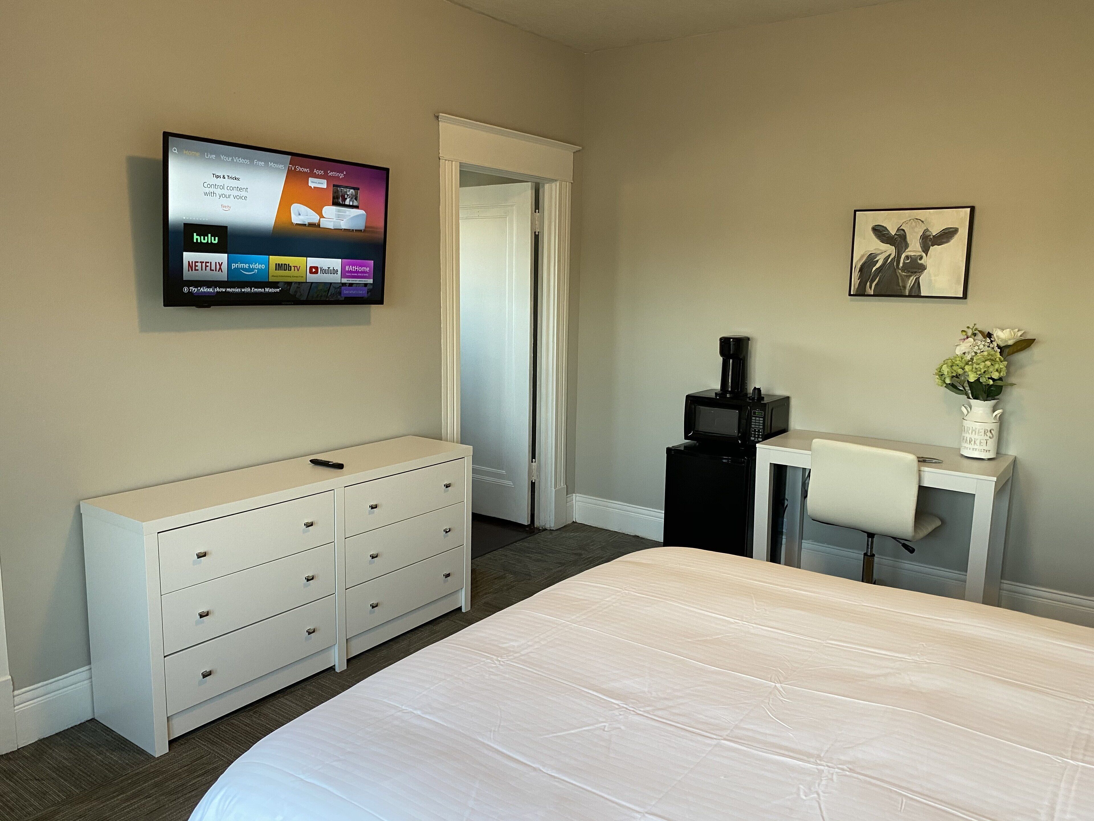 A hotel room with a bed, a dresser, a desk with a chair, a microwave, a television mounted on the wall, and a framed picture on the wall.