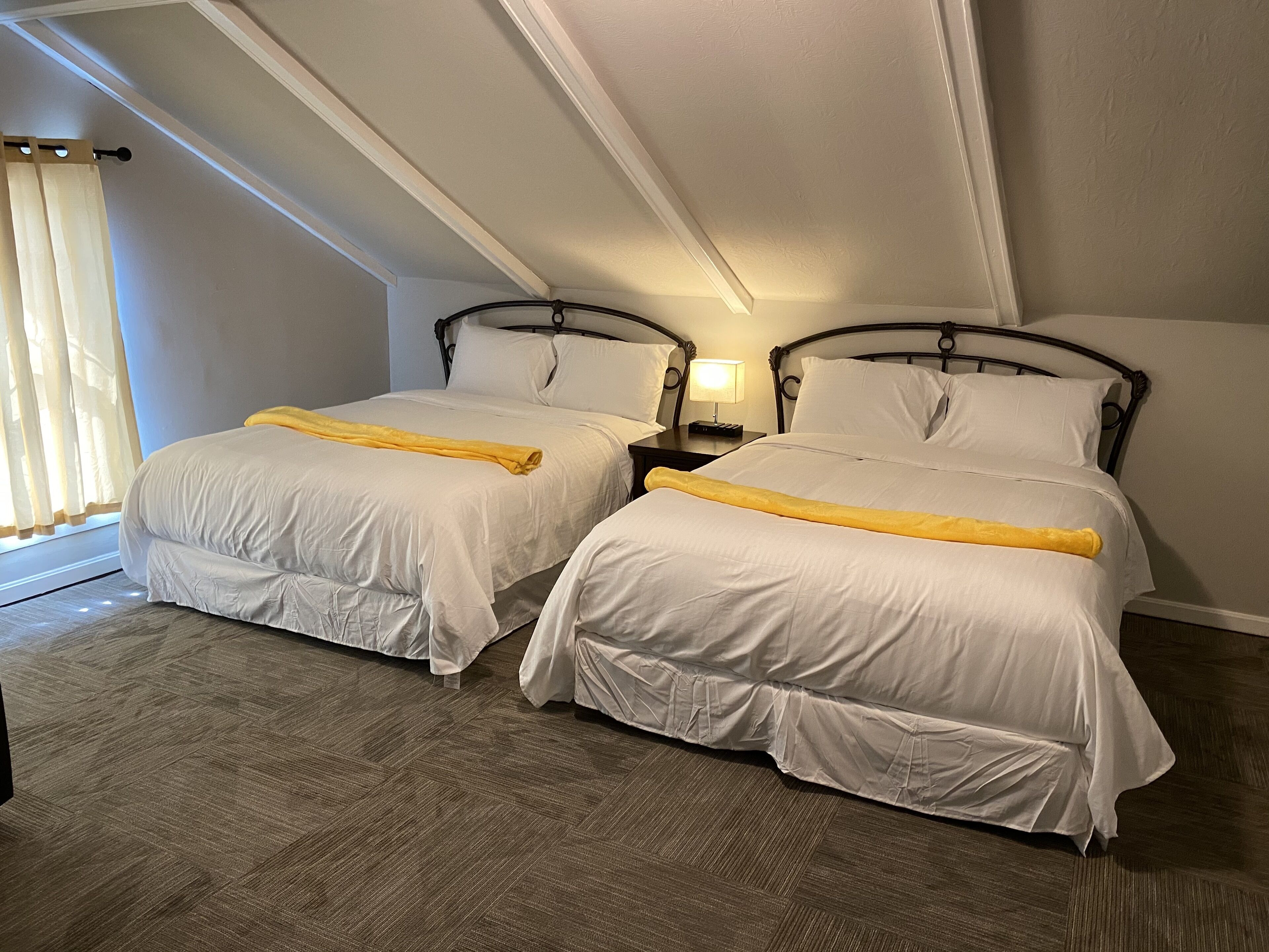 A hotel room with two beds, a nightstand with a lamp, and a window with curtains.