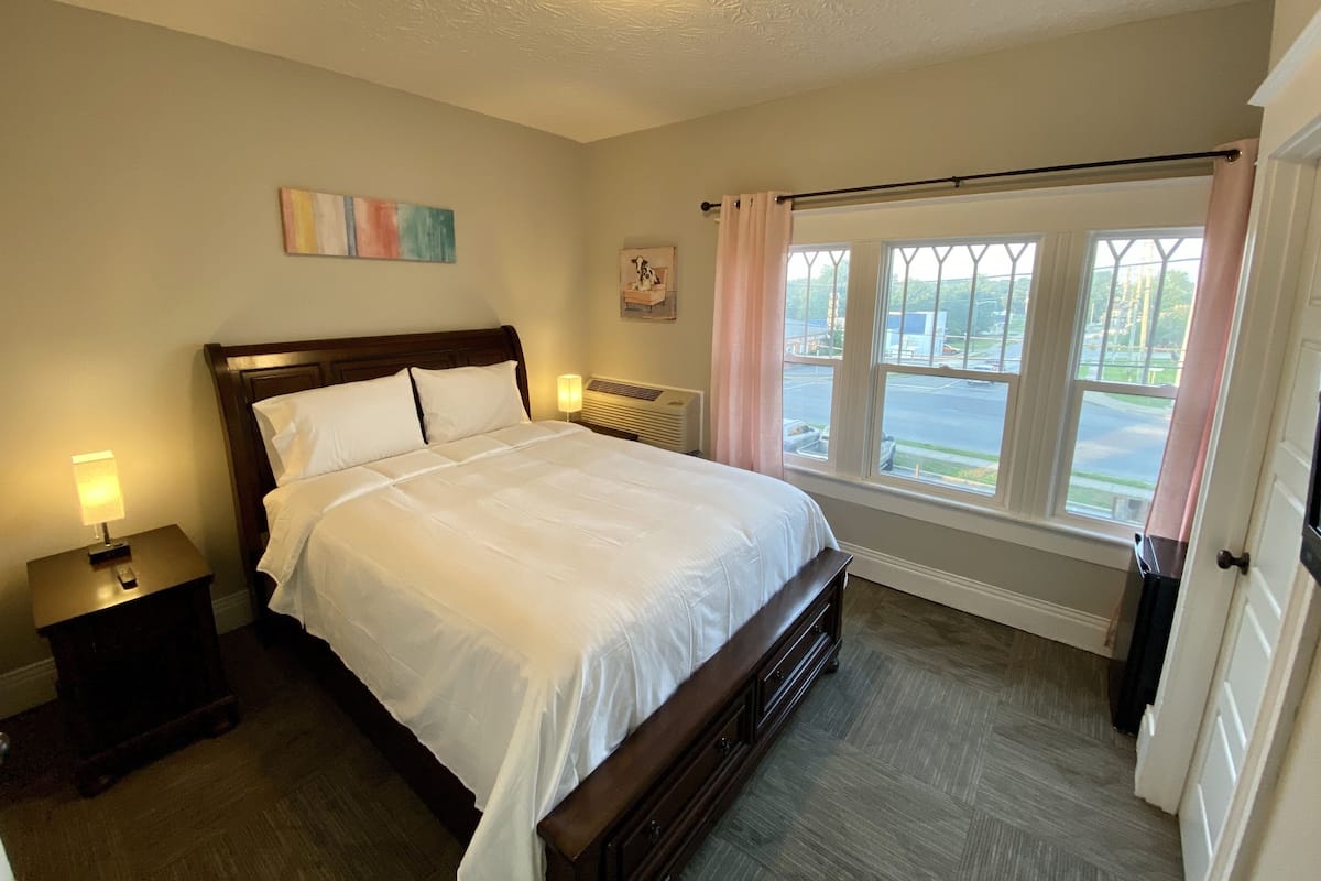 A neatly made bed in a room with a large window, a radiator, and a painting on the wall.